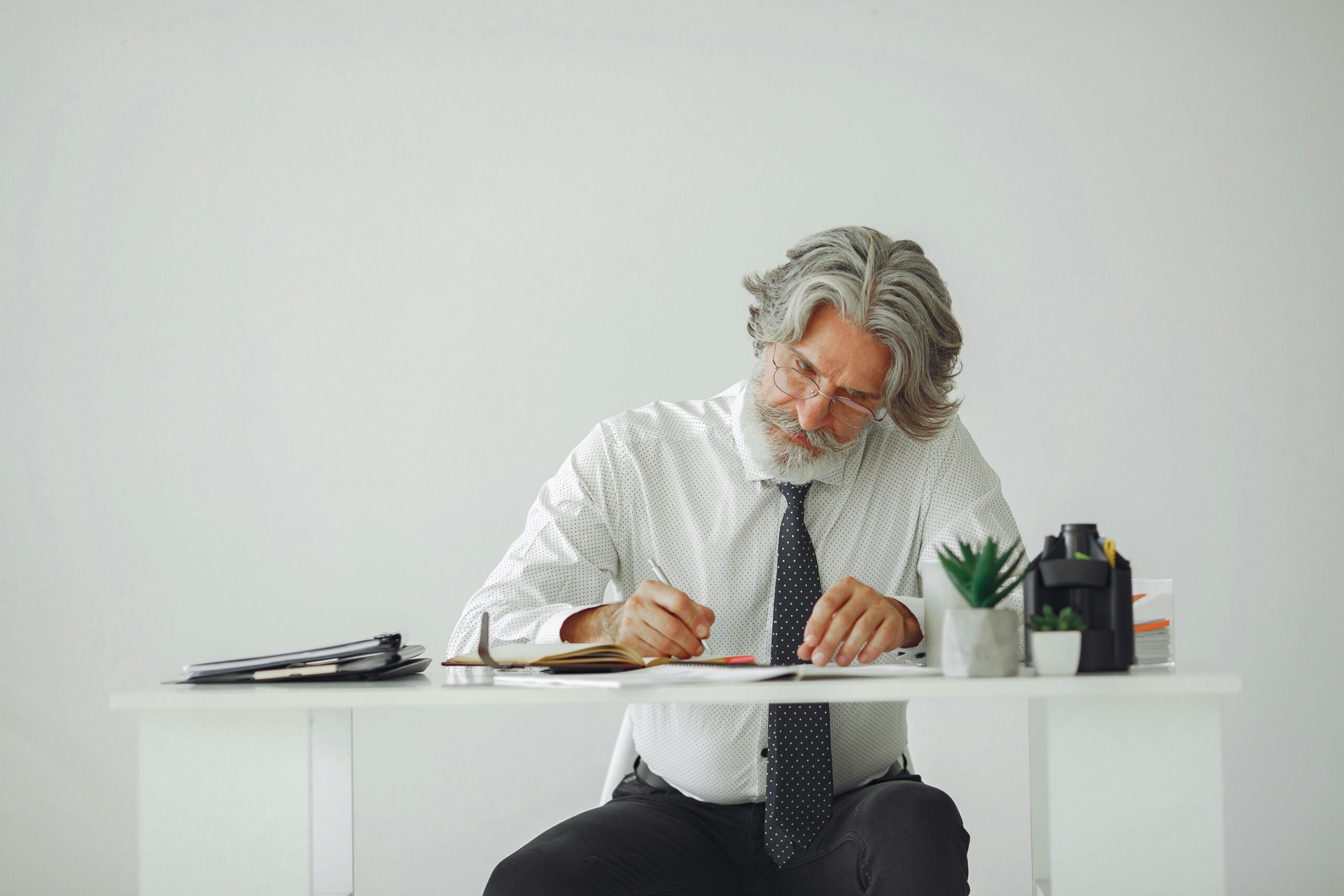 Elderly businessman with gray hair writes at a desk in a modern office.
